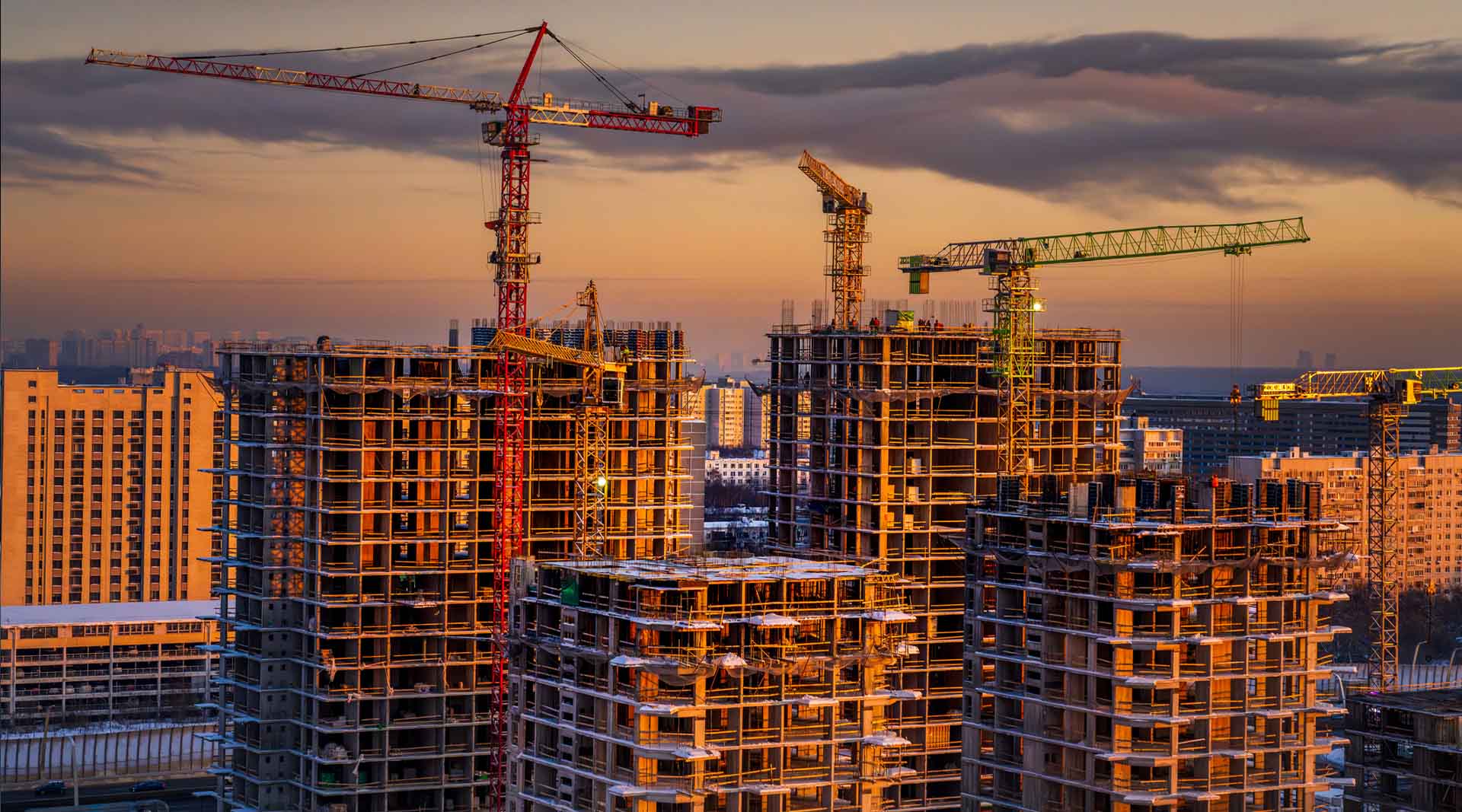 Large construction site with tower cranes and concrete high-rises under a dramatic golden sunset sky.