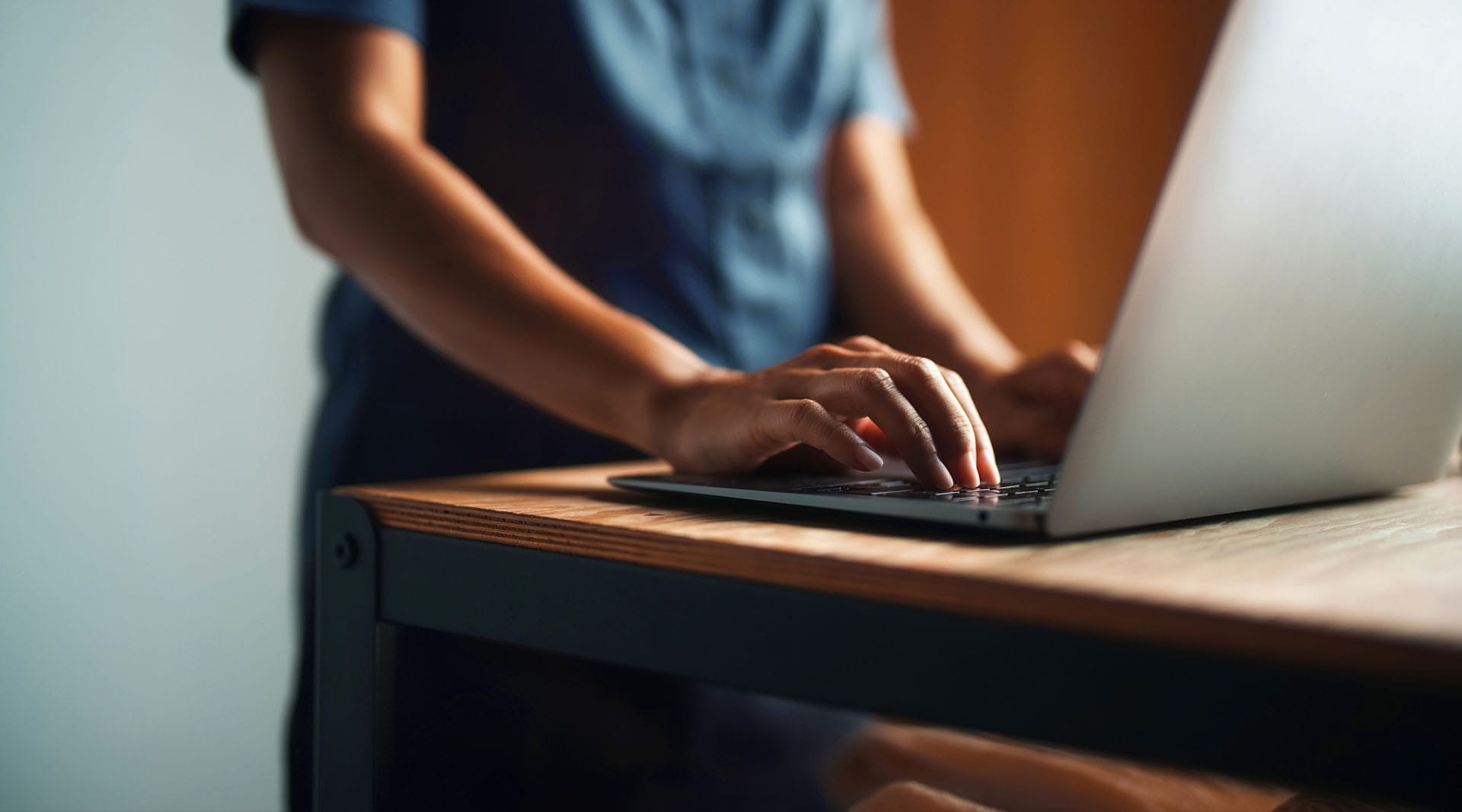 Person typing on a silver laptop at a wooden standing desk in soft warm lighting.